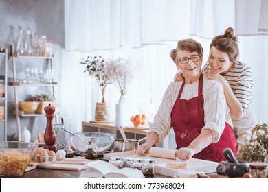 Smiling senior woman in red kitchen apron rolling out dough and granddaughter standing behind her - Powered by Shutterstock