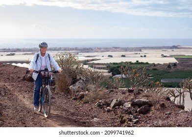 Smiling Senior Woman Riding In A Country Road With Her Bike, Cyclist Wearing Sport Helmet And Backpack. Horizon Over Water