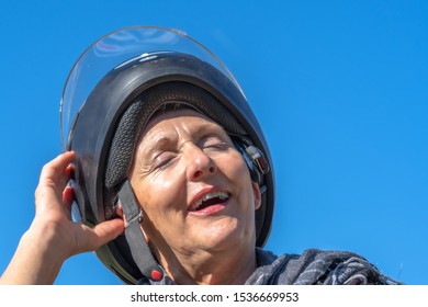 Smiling Senior Woman With Scarf On Her Neck Putting On Black Motorcycle Helmet With Blue Sky In Background