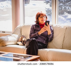 Smiling Senior Woman Sitting On Sofa By Large Window Looking At Her Phone On Sunny Winter Day; Snow Outside The House