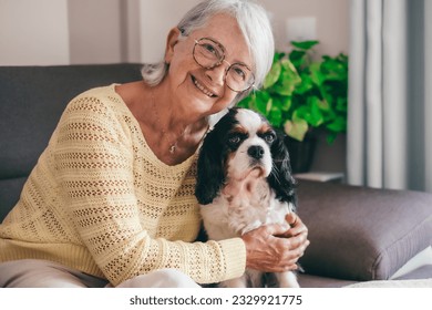 Smiling senior woman sitting on sofa at home while hugging her cavalier king charles spaniel dog. Elderly retired lady with her best friend looks into the camera - Powered by Shutterstock