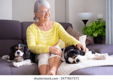 Smiling senior woman sitting on sofa with her two Cavalier King Charles dogs cuddling close up. Relaxing time of her spending quality time with her beloved pets beside her - Powered by Shutterstock