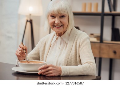 smiling senior woman sitting at table and eating cream soup at home - Powered by Shutterstock