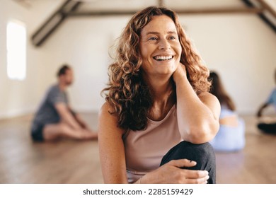 Smiling senior woman sitting in a yoga studio with a group of people exercising in the background. Happy mature woman having a workout session with her class in a community fitness center. - Powered by Shutterstock