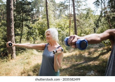 Smiling senior woman training with dumbbells near african american husband in forest