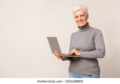 Smiling Senior Woman Using Laptop, Browsing Internet While Standing Against Light Studio Background. Copy Space