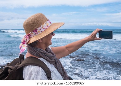 A Smiling Senior Woman In Vacation At The Sea Takes A Photo Of The Horizon Over Water. Casual Clothing With Backpack And Hat. Cloudy Sky