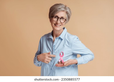 Smiling senior woman wearing eyeglasses showing pink ribbon isolated on beige background. Health care, support, prevention. Breast cancer awareness month concept - Powered by Shutterstock