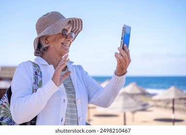 Smiling senior woman wearing hat and sunglasses standing at the beach using mobile phone in video chat, horizon over sea and blue sky - Powered by Shutterstock