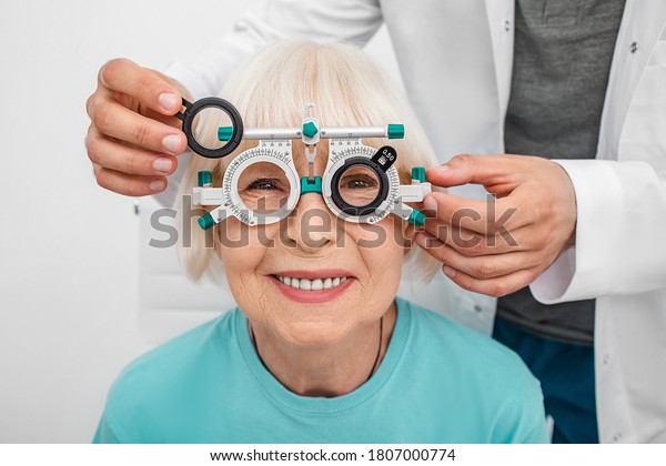 Smiling senior woman wearing\
optometrist trial frame at ophthalmology clinic. Ophthalmologist\
helping select glasses for treatment of vision. Eye\
check-up