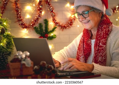 Smiling Senior Woman Wearing Santa Hat At Christmas Time Typing On Laptop From Home Getting In Touch With Family Or Friends