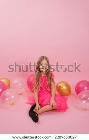 Smiling seven-year-old girl dressed in a fashionable dress sits on a pink background surrounded by balloons. Celebrate your birthday