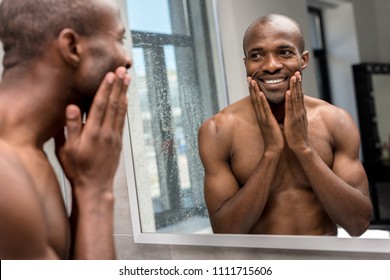 Smiling Shirtless African American Man Applying Shaving Lotion And Looking At Mirror