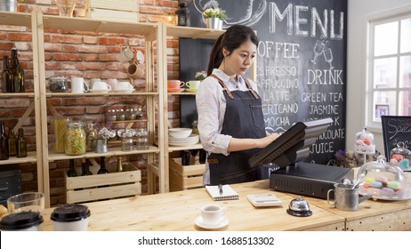 smiling shop assistant using pos point of sale terminal to put in order from note at restaurant register. young girl waitress standing in wooden bar counter and working on digital tablet in cafe - Powered by Shutterstock