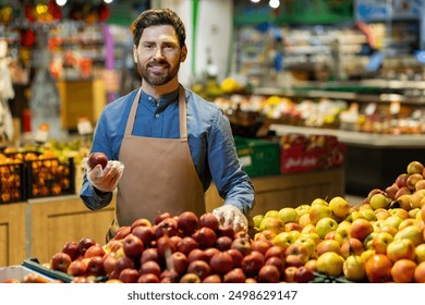 Smiling shopkeeper holding apple at fruit stand in grocery store. Man in apron displaying fresh produce, engaging with customers, promoting healthy eating and organic food - Powered by Shutterstock