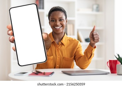 Smiling short-haired young black woman in yellow shirt sitting at workdesk with laptop, freelancer showing modern smartphone with white empty screen and thumb up, mockup, copy space