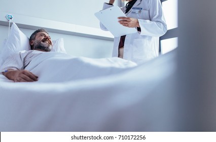 Smiling Sick Man Lying In Bed With Doctor Standing By. Nurse Visiting Male Patient In Hospital Ward.