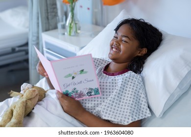 Smiling Sick Mixed Race Girl Lying In Hospital Bed Reading Get Well Greetings Card And Holding Teddy. Medicine, Health And Healthcare Services.