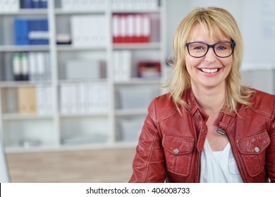 Smiling Single Blond Business Woman In Red Leather Jacket And Eyeglasses At Small Office With Copy Space Over Bookshelf Area