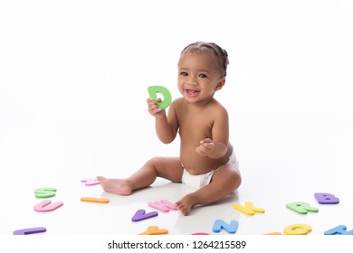 A smiling six month old baby girl wearing a white diaper. She is sitting on a white seamless background and playing with foam alphabet toys.