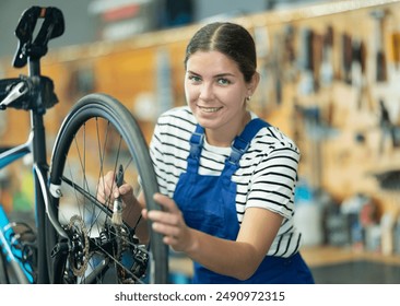 Smiling skilled young female mechanic in blue overalls adjusting bicycle wheel, cleaning gears with small brush in professional bike repair shop - Powered by Shutterstock