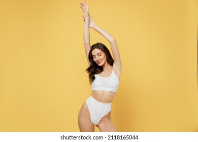 Smiling slim beautiful lovely attractive young brunette woman 20s in white underwear with perfect fit body standing posing raise up hands close eyes isolated on plain yellow background studio portrait