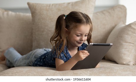 Smiling Small 7s Caucasian Girl Relax On Sofa In Living Room Look At Pad Screen Talk On Video Call Online. Happy Cute Kid Lying On Couch At Home Use Tablet Play Or Study Distant. Technology Concept.