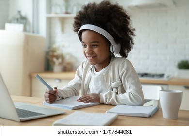 Smiling Small African American Girl In Headphones Watch Video Lesson On Computer In Kitchen, Happy Little Biracial Child In Earphones Have Online Web Class Using Laptop At Home, Homeschooling Concept