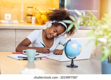 Smiling Small African American Girl In Headphones Watch Video Lesson On Computer In Kitchen, Happy Little Biracial Child In Earphones Have Online Web Class Using Laptop At Home, Homeschooling Concept