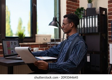 Smiling Small Business Owner Looking At Laptop Screen With Analytics Data Holding Clipboard Sitting At Desk. Entrepreneur With Glasses Comparing Business Charts Happy With Results.