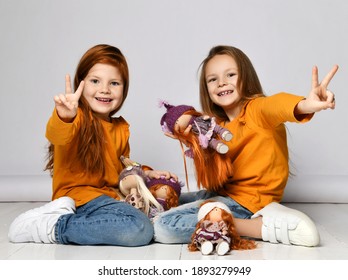 Smiling small girls sisters in yellow comfortable longsleeves, jeans and sneakers sitting on floor playing with toy dolls and showing peace signs over white background. Happy childhood, stylish look