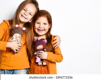 Smiling small pretty girls sisters or friends in terracotta yellow comfortable longsleeves standing hugging holding toy dolls in hands over white background, copy space. Happy childhood, stylish look