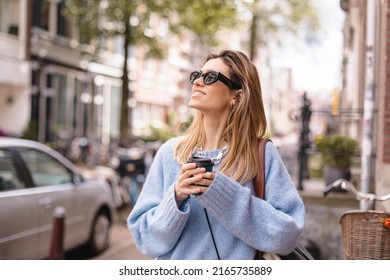 Smiling smart blonde haired woman holding a paper cup of coffee take away outdoors. Woman wear black sunglasses and blue sweater. Happy smiling woman walk outdoor on the street. Girl look upper corner - Shutterstock ID 2165735889