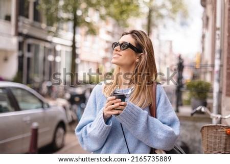 Smiling smart blonde haired woman holding a paper cup of coffee take away outdoors. Woman wear black sunglasses and blue sweater. Happy smiling woman walk outdoor on the street. Girl look upper corner
