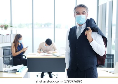 Smiling And Smart Caucasian Businessman In Black Suit And Asian Teamwork Wearing Masks Prevent Covid 19 Virus At Office. New Nomal And Social Distace Of Business Concept.