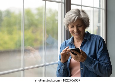 Smiling smart elderly grey-haired 60s woman have fun texting or messaging on modern smartphone gadget at home. Happy mature Caucasian female laugh browsing internet or surfing on cellphone device.
