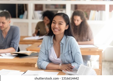 Smiling Smart Female Asian Teen Girl Looking At Camera Sitting At Desk Studying In Classroom With Diverse Classmates Group, Happy Chinese College University Student In International Class Portrait