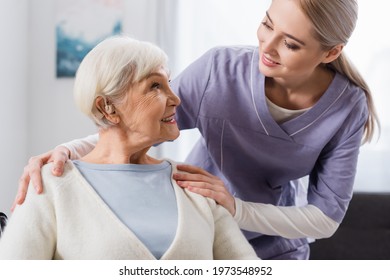 Smiling Social Worker Hugging Shoulder Of Senior Woman With Hearing Aid