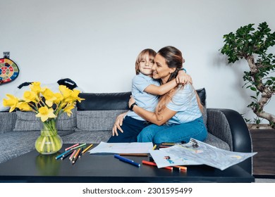 Smiling son hugging his happy mother during home family weekend Mother   son drawing and color pencils   markers in the living room  children creativity concept  Selective focus 