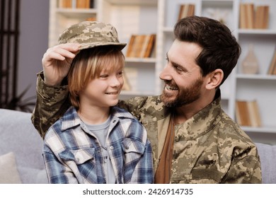 Smiling son wearing his father's army hat, sits on his dad's knees. Father, dressed in Ukrainian military gear, delighted to be back home, aims to nurture a patriotic spirit in his child - Powered by Shutterstock