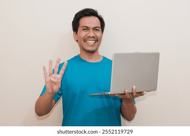 Smiling Southeast Asian man wearing green t-shirt casual clothes holding laptop with giving number 12345 by hand gesture, standing over isolated white background, Positive person - Powered by Shutterstock