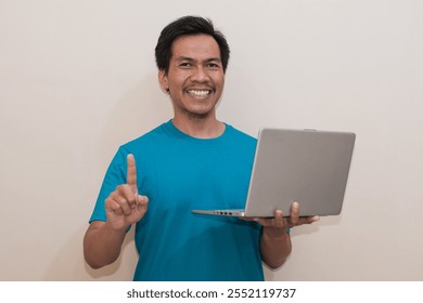 Smiling Southeast Asian man wearing green t-shirt casual clothes holding laptop with giving number 12345 by hand gesture, standing over isolated white background, Positive person - Powered by Shutterstock