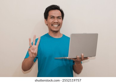 Smiling Southeast Asian man wearing green t-shirt casual clothes holding laptop with giving number 12345 by hand gesture, standing over isolated white background, Positive person - Powered by Shutterstock