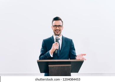 Smiling Speaker Gesturing And Talking Into Microphone At Podium Tribune During Seminar In Conference Hall