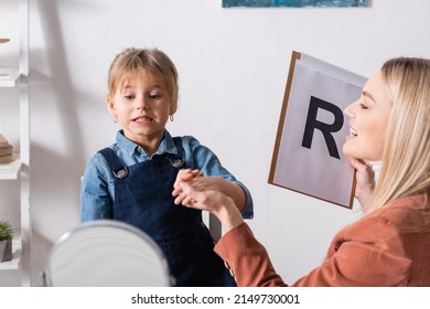 Smiling Speech Therapist Holding Clipboard And Hand Of Child Near Mirror In Consulting Room