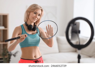 Smiling sports blogger holding badminton racket while streaming online fitness lesson with smartphone at home - Powered by Shutterstock