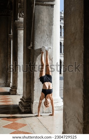 Smiling sportswoman in black shorts and crop top doing handstand near column of building on street in Venice