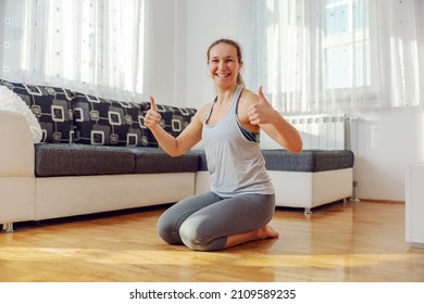 Smiling sportswoman kneeling on the floor at home and showing thumbs up. Even it is corona, you can have healthy life.