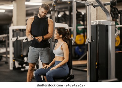 Smiling sportswoman sitting at chest machine and looking at tablet while having consultations with her coach at gym. - Powered by Shutterstock