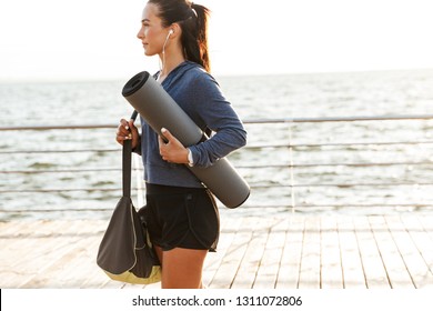 Smiling sportswoman walking at the beach, carrying fitness mat - Powered by Shutterstock